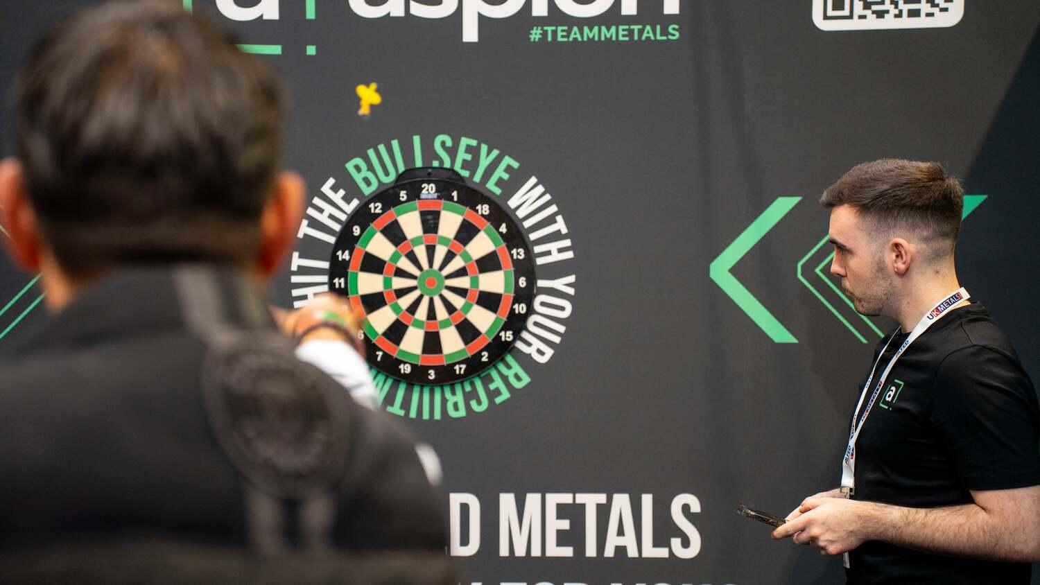 Aspion Search team member overseeing a darts game at the UK Metals Expo stand, with a branded backdrop reading "Hit the Bullseye with Your Recruitment".        Ask ChatGPT